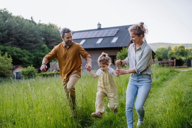 En familj på tre går hand i hand i ett gräsbevuxet fält nära ett hus med solpaneler.