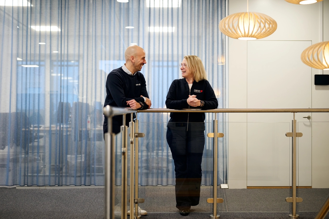 Två kollegor står lutade mot ett räcke i en modern kontorsmiljö och samtalar med varandra.