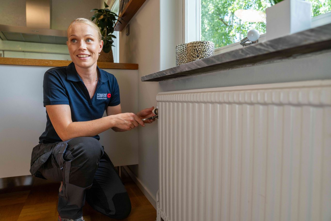En person knäböjer bredvid en vit kylare och justerar en ventil. De är klädda i blå pikétröja och gråa byxor i ett väl upplyst rum med synligt fönster och grönska utanför.